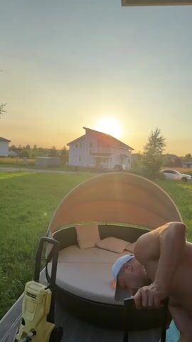 The video opens with a man wearing a white baseball cap and black and white patterned shorts lying on a large, round outdoor lounge chair. He is partially obscured by the chair's canopy and is facing downwards, with his arm extended. The sun is setting in the background, casting a warm glow over a white house and green lawn. The scene then shifts to show white curtains hanging in an outdoor covered area. The camera pans to reveal a table with a grill basket, food, drinks, and various objects. A woman in a white t-shirt and shorts is sitting on a grey sofa, holding a microphone and speaking. The camera then pans across the sofa and out towards a window, showing a glimpse of what appears to be exercise equipment and construction materials in the background. The video ends with a shot of the grey sofa with pillows, a remote control, and a pink smartphone on it. The overall environment is an outdoor living space that appears to be a patio or deck attached to a house.