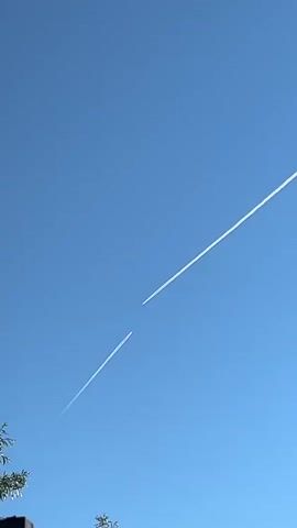 The video shows a clear blue sky with two contrails from airplanes. The contrails are white and stretch across the frame from the bottom left to the top right. The lower contrail is longer and appears to be fading into the distance, while the upper contrail is shorter and is behind the lower contrail. The two contrails are separated by a small gap. There is a small portion of a dark structure and some green foliage visible at the bottom of the frame, suggesting the vantage point is from the ground. The lighting indicates it is daytime, likely morning or afternoon.