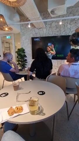 The video captures an indoor event, likely a presentation or talk, in a modern, well-lit space. The setting resembles a cafe, conference room, or a contemporary office common area, featuring light-colored tables and chairs, decorative woven ceiling lamps, and a stone-textured accent wall.

The central focus is a large screen displaying "Deloitte AI in action," with additional Hebrew text partially visible, including "מפגש" (meeting). A woman stands at the front, addressing the attendees, suggesting she is the speaker.

Numerous people, both men and women, are present. Some are seated at tables in the foreground, enjoying drinks (coffee, clear beverages) and snacks (pastries like croissants). Others are standing or sitting further back, observing the speaker. Several individuals are seen holding smartphones, possibly taking photos or videos. Towards the end of the clip, some attendees are seen clapping, indicating appreciation or the conclusion of a segment. The overall atmosphere appears engaged and relaxed. The event seems to be a professional gathering centered around AI, hosted by Deloitte. While the user provided "Al Jizah, Jordan" as context, the presence of Hebrew text on the screen is a notable visual detail.