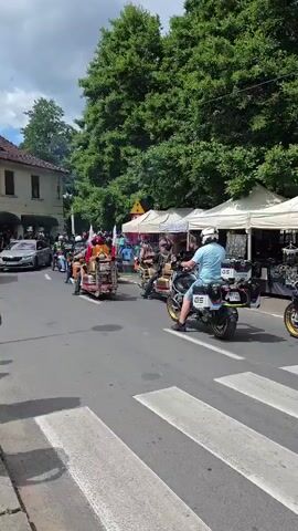 A group of motorcyclists are riding in a procession down a street. They are passing by tents set up on the side of the road, likely for a market or event. A red food truck with the words "ZABIEX FRYTKI BELGIJSKIE" on it is parked on the right side of the street. A pedestrian is standing near the food truck. In the foreground, a crosswalk is visible. Several cars are parked on the left side of the street. The street is lined with trees. The weather appears to be sunny.