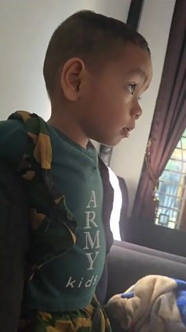 A young boy with dark hair is looking away from the camera. He is wearing a green t-shirt with "ARMY kids" printed on it in white letters, and camouflage pants. He is standing in front of a window with dark curtains. To the right of the boy, there is a couch with a blue and white patterned blanket on it. Further back, an open doorway reveals an outdoor area with sunlight. A broom with a yellow handle is leaning against the wall near the doorway. A television is mounted on the wall, displaying a video of hands playing with a toy ice cream set. Below the television are two large speakers and a vase with orange flowers. A blue inflatable toy animal and an orange ball are on the floor. A baby bottle is on top of one of the speakers.