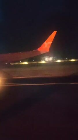 The video shows an airplane's wing with an orange fin and a white logo in the night sky. The airplane is in the process of taking off or landing, as evidenced by the runway lights visible below and the blur of motion. The surrounding environment appears to be an airport at night, with a dark sky and illuminated runway.