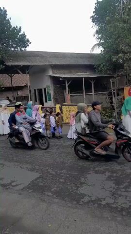 A group of people, most of whom are wearing white clothing and headscarves, are gathered on a paved road. Some are walking in a line, while others are standing and talking. Several motorcycles and cars are present, suggesting a procession or event. The environment appears to be a rural or suburban area with trees and buildings in the background. The atmosphere seems festive or celebratory.