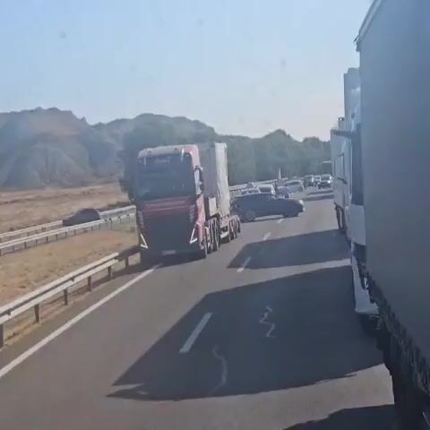 A red semi-truck with a white trailer is stopped on a highway. To the right of the red semi-truck is a white semi-truck. To the left of the red semi-truck, there is a dark-colored car that appears to have its front end turned toward the right lane. In the distance, there are more vehicles stopped on the highway, and people are visible standing outside of their vehicles. The highway is bordered by a guardrail on the left. The landscape is dry and hilly with sparse vegetation. The sky is clear and blue. The sun is shining.