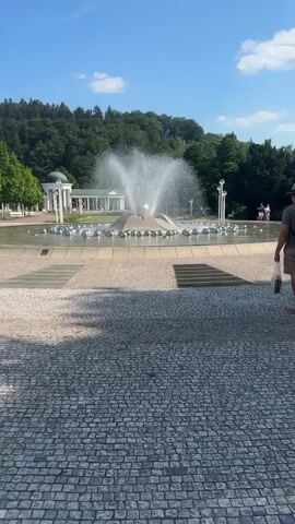 A video shows a large fountain in a public square. The fountain features water spraying upwards from a central structure, with decorative spheres arranged around the base. In the background, there is a white colonnaded building with a dome, surrounded by trees and a hill. The ground in the foreground is paved with cobblestones and square tiles. The sky is blue with a few scattered clouds. There are a few people visible in the background walking near the building. The overall scene appears to be a park or a plaza on a sunny day.