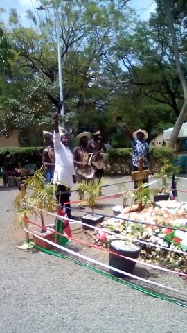 The video depicts an outdoor ceremonial gathering in Bondo, Kenya, taking place during bright daylight. Several people are present, clustered around a central floral display.

In the foreground, a person wearing a white shirt and a feathered headpiece stands, facing to the right. Behind them, another individual in a similar white shirt and headwear is visible. Further into the scene, a group of people are seated or crouching; one appears to be playing a percussion instrument. To the right, a person in a patterned blue shirt and a straw hat stands near the main display.

The focal point of the scene is an elaborate arrangement of fresh flowers in white, red, and green, possibly situated on a small mound or platform. Small Kenyan flags are interspersed within the flowers, and red, white, and green ribbons are strung across the area, creating a decorative boundary. Several potted plants also contribute to the decor.

The background features large, leafy trees and a paved road or path in the immediate vicinity. A building with blue sections can be seen in the distance. The overall atmosphere suggests a cultural event or a memorial service, marked by the traditional attire, the presence of music, and the national colors incorporated into the decorations. The audio includes a distinct vocalization or chant. No discernible text is visible within the frame.