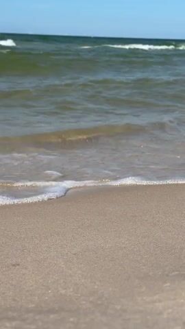 The video shows a close-up view of gentle waves lapping onto a sandy beach. The water is a murky green, and the sky is a clear blue. The focus is on the sand and the water's edge, with the waves creating small white foam as they recede. The scene is peaceful and serene, capturing the natural beauty of the coastline.