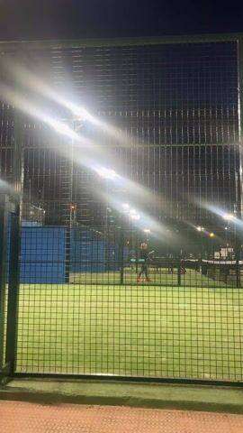 The video shows a padel court at night. The court is surrounded by a fence of green metal bars, and is lit up by several bright, overhead lights. Two people can be seen inside the court. The person closest to the camera is wearing dark clothes and red shoes. They are holding a padel racket and appear to be hitting a ball. The other person is further away and harder to see clearly. The floor outside the court is paved with brown and red rectangular blocks.