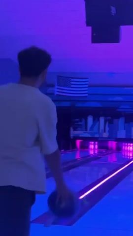 A person in a white t-shirt and dark pants is bowling in a dimly lit bowling alley. The lanes are illuminated with purple and blue neon lights, and the pins at the end of the lane are lit up in green and purple. An American flag is visible on the wall in the background. The bowler releases the ball and then walks back towards the camera.