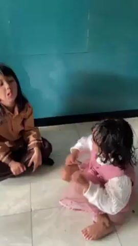 Two young girls are seated on a tiled floor in front of a blue wall. The girl on the left wears a brown uniform and sits with her legs crossed. She looks at the camera briefly and then glances at the girl on the right. The girl on the right wears a pink dress with a white undershirt and her hair appears wet. She is initially crawling and then sits up, looking towards the camera. The girl in brown gives a thumbs up.