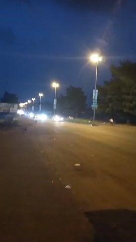 The video depicts a nighttime street scene. Several cars are driving on a paved road illuminated by streetlights. The sky is a dark blue, suggesting it is late evening or night. Trees line the side of the road, appearing dark against the night sky. The camera appears to be in a moving car, capturing the passing scenery. There are glimpses of buildings in the background. The overall atmosphere is that of a typical night drive in a suburban or urban area.