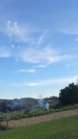 The video shows a rural landscape under a partly cloudy blue sky. In the foreground is a field of tall grass. Behind that is a vineyard and further in the background are trees, a small number of buildings, and low hills. Two plumes of gray smoke are rising from the direction of the buildings. Several white streaks, likely from fireworks, are visible in the sky. The sound of fireworks going off can be heard in the background.