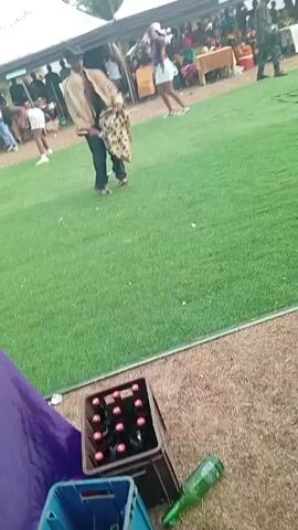 The video shows a man dancing on a green lawn at what appears to be an outdoor event. There are white tents set up in the background, with people gathered under them. The man is wearing a hat, a brown shirt, and dark pants, and he is carrying a bag with a busy pattern. He is performing a dance that involves kicking his legs. Other people, including women in dresses, are visible in the background. A case of drinks is visible in the foreground.