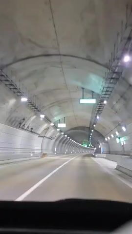 The video shows the view from inside a vehicle driving through a long, modern tunnel. The tunnel has a high, arched ceiling made of concrete sections. The walls are also concrete, with a ribbed texture. The tunnel is illuminated by rows of bright lights spaced along both sides of the ceiling. There are also green illuminated signs hanging from the ceiling at intervals. The road is paved and has a white lane divider. The interior of the vehicle's dashboard is partially visible at the bottom of the frame. The tunnel appears to be empty of other vehicles.