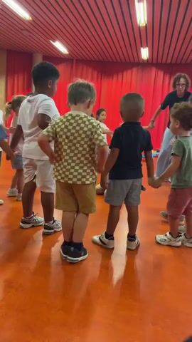 The video shows a group of young children, about 5 to 7 years old, holding hands and dancing in a circle on an orange floor in a room with red curtains and a slatted ceiling. A woman in the background is watching them. The children are wearing casual clothes and sneakers. The room appears to be a classroom or a community hall.