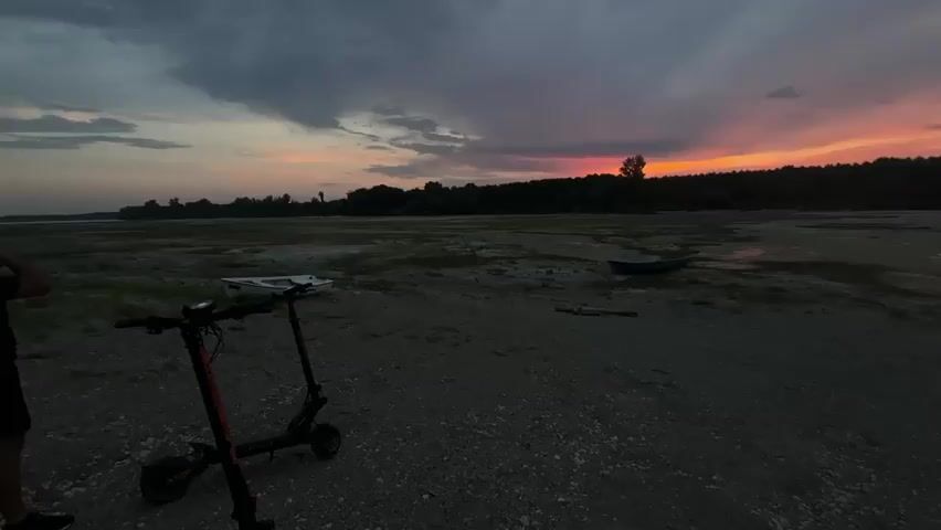 The video captures a low-angle, wide shot of a desolate, muddy landscape at dusk. The sky is a dramatic canvas of dark, swirling clouds streaked with vibrant hues of pink, orange, and yellow from a setting sun. A silhouetted treeline forms the horizon, with a single, sparse tree standing out in the middle. In the foreground, a black and orange electric scooter is parked. To the left of the scooter, a person's legs and lower torso are visible, their body angled away from the camera. Further into the scene, a small, dark blue boat rests on the mud, and a single stick lies on the ground nearby. The ground itself is a mix of dirt, small pebbles, and patches of shallow water reflecting the colorful sky. The overall atmosphere is one of quiet solitude and the fading light of day.