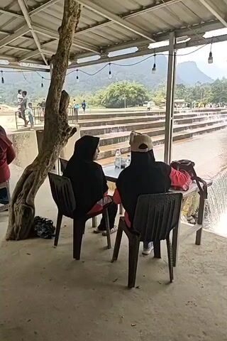 The video shows two individuals seated at a table under a covered outdoor structure. One person, wearing a black headscarf and a red shirt, is seated on the left. The other person, wearing a black headscarf and a tan cap, is seated on the right. A table with a white surface is between them, with a bottle of water on it. In the background, a stepped water feature or cascade is visible, with water flowing over the steps. Beyond the water feature, there is a rocky riverbed, trees, and mountains in the distance. Other people can be seen in the background, including children playing near the water. The camera pans to the right, showing more of the river and the surrounding landscape. A person in a light-colored striped shirt is then seen standing to the right of the frame, looking towards the left. The camera then pans to the right again, showing a man in a blue shirt standing and looking downwards. Another table with a white surface is in the foreground. Further to the right, more people are seated under the covered structure, including a person wearing a light blue headscarf and a person wearing a white headscarf. The video appears to be a personal recording taken at a scenic outdoor location, possibly a resort or park.