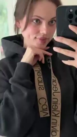 A fair-skinned woman wearing a black Calvin Klein hoodie poses for a selfie. She has brown hair pulled back, green eyes, and dimples, and smiles at the camera. The background is a room with white walls and a door.  The woman holds a black smartphone in her right hand to take the photo.