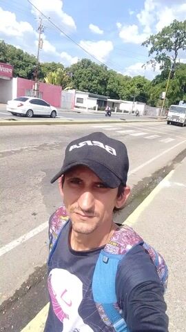A man is filming a selfie video standing on a sidewalk near a road. He is wearing a black baseball cap with the word "BEBA" on it, a dark t-shirt, and a backpack with a colorful pattern and blue straps. He has short dark hair and a light beard. He is looking at the camera with a serious expression. 

Behind him, there is a road with cars and trucks passing by. A white truck with a flatbed loaded with wooden planks drives past. There are trees and buildings on either side of the road. The sky is blue with some clouds. The sun is shining.