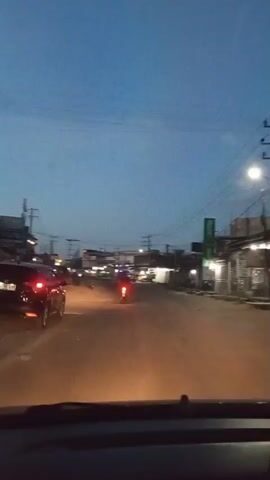 The video shows a street scene at dusk, viewed from inside a car. The street is lined with buildings and power lines are visible overhead against a blue sky. The car's headlights illuminate the road ahead. A car and a motorcycle are also visible in the distance. A child's voice can be heard in the background.