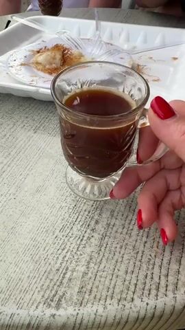 A woman’s hand with red nails holds a small glass cup of dark brown liquid, likely coffee, at an outdoor table. Other people are partially visible in the background, sitting at the same table. A white styrofoam plate with leftover shredded pastry is also on the table. The table appears to be made of wood and is painted in white and grey. The focus is on the cup and the woman's hand.