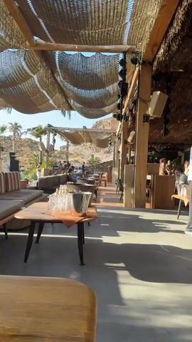 The video pans across an outdoor lounge area, likely a beach club or restaurant. The scene is dominated by a woven canopy providing shade, supported by wooden beams. In the foreground, a low wooden table holds several glasses and a metal ice bucket. Further back, comfortable seating areas with cushioned sofas are visible. People are scattered throughout the space, some seated, others standing and interacting. Palm trees and rocky hills form the backdrop, suggesting a warm, arid climate. The atmosphere appears relaxed and social, with what seems to be a DJ setup or sound system visible in the mid-ground, indicating potential music or entertainment. The overall impression is a stylish, open-air venue designed for leisure and socializing.