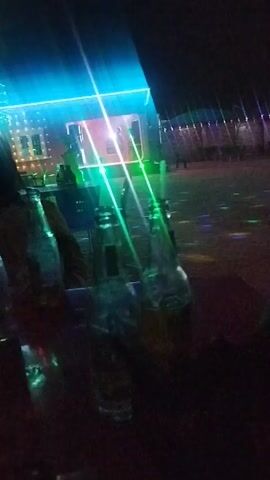 The video shows a bar scene at night. Several empty glass beer bottles are on a table, and a person is holding a smartphone, likely taking a photo or video. The background is dimly lit with blue, green, and pink lighting effects. People can be seen in the background, suggesting an active social atmosphere. The overall ambiance indicates a festive or party setting.
