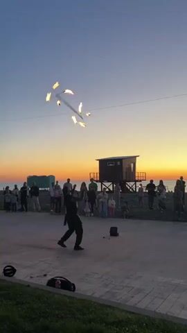 Against the backdrop of a sunset beach, a fire performer entertains a crowd. The performer, dressed in black clothing, skillfully manipulates burning torches, creating a dazzling display of flames. A lifeguard tower stands in the background, and onlookers of all ages watch the performance with interest. The performance takes place on a brick-paved area near the beach, with audio equipment nearby. The scene is bathed in the warm glow of the setting sun.