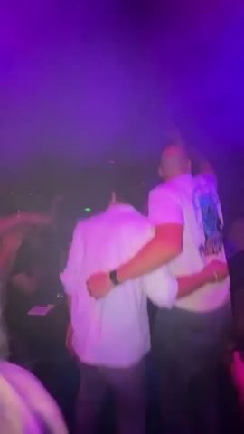 A group of young adults are in a dimly lit nightclub. Two men in the foreground are facing away from the camera. The man on the left is wearing a white short-sleeved button-down shirt, and the man on the right is wearing a white t-shirt with a graphic on the back. The man on the right has his arm around the man on the left's shoulder. Both men appear to be dancing or moving to music. The background is filled with a crowd of people, many of whom are also dancing or raising their arms. The room is illuminated by colored lights, primarily pink and purple, and there is visible smoke or haze in the air. Some people in the background appear to be holding drinks.
