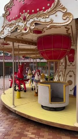 The video shows a stationary carousel. The carousel features a red-and-white ornate top, gold decorative accents, a yellow floor, and various figures including horses of different colors, a hot air balloon shaped seat, and a red toy car. It is located outdoors on a brick surface.
