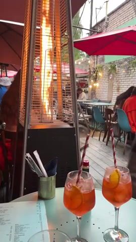 The video shows an outdoor seating area of a restaurant with a patio heater in the foreground. Two orange cocktails with straws and orange slices are on a table. A menu is also visible on the table. In the background, people are seated at tables, and there are red umbrellas and string lights. A person wearing a black t-shirt with a graphic print and denim shorts is walking away from the camera. Another person with long brown hair is seated to the left. The patio heater has flames visible through its protective grate. The environment appears to be a casual dining setting, possibly in the evening or on a cooler day due to the presence of the heater.