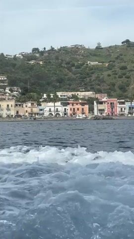 The video captures a scenic view of a coastal town, likely Lipari, Italy, from the perspective of a moving boat. In the foreground, foamy white and blue water, a wake created by the boat, churns across the bottom of the frame. Beyond the water, a line of colorful buildings, including houses in shades of white, beige, orange, and pink, stretches along the shoreline. These structures are nestled at the base of lush green hills and mountains that rise steeply in the background, dotted with more buildings and vegetation. The sky above is bright and clear, suggesting a daytime scene with pleasant weather. No people or visible text are discernible in the video.