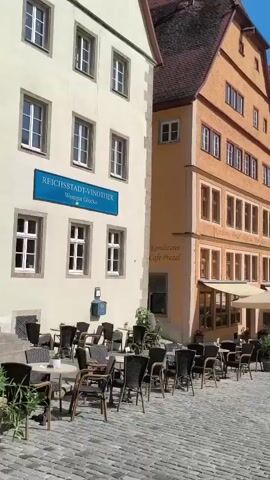 The video pans across a European town square. Several people are seated at outdoor tables in front of buildings with historic architecture. The buildings feature colorful facades and steeply pitched roofs. A cobblestone street leads into the distance. The sky is blue with some clouds.