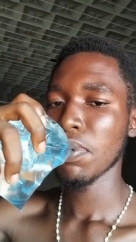 A shirtless, dark-skinned man with short black hair, a beard, and a white beaded necklace is drinking water from a clear plastic bag. He is looking directly at the camera while holding the bag to his lips and tilting his head back. The background consists of a ceiling with a grid-like pattern.