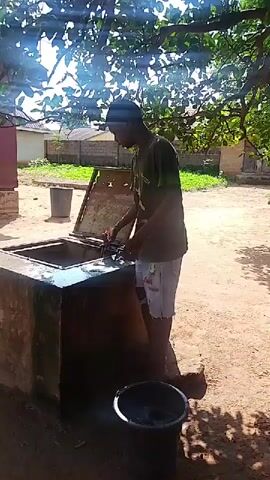 A young person is standing on a stone structure next to an open well. The person is wearing a dark t-shirt and light-colored shorts with a pattern. They are using a rope to lower a bucket into the well. Sunlight filters through the leaves of a tree overhead, casting shadows on the ground. In the background, there are buildings and a large plastic bin.