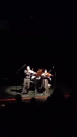 A darkened theater room shows a stage with two violinists and a piano. Both musicians are dressed in dark formal wear. A microphone is positioned in front of each performer. The light on stage is focused on the musicians while the audience is silhouetted in the darkness. The two violinists are playing.