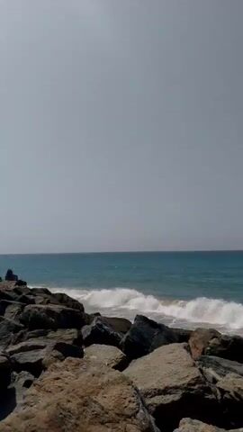 The video shows a beach scene, with waves gently lapping against the shore. In the foreground are large rocks that seem to form a barrier between the viewer and the water. The ocean is a tranquil turquoise color.  The sky is overcast with a pale grey, suggesting a cloudy day.