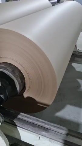The video shows a large roll of light brown paper being unwound by a machine. The roll is cylindrical and tightly wound, sitting on a metal cylinder that is part of the machine. The machine has metal bars and rollers that support the paper roll and guide it as it unwinds. The background is a dimly lit industrial setting. The camera remains static throughout the video.