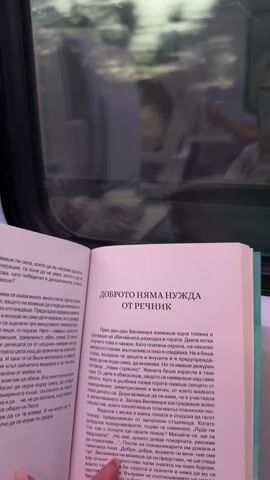 A medium shot captures a person reading a book on a train. The book, open to a Bulgarian text titled "ДОБРОТО НЯМА НУЖДА ОТ РЕЧНИК" (Good doesn’t need a dictionary), takes up most of the frame. The pages are white with clear black text. The person's hand, holding the book, is visible on the left side. 

Through the train window, blurred and out of focus, a landscape transitions from day to dusk. The sky is a mix of fading daylight and the early colors of sunset. Hints of trees and fields are visible, indicating the train is moving through a rural area. The scene is illuminated by natural light, and the overall mood is calm and introspective.