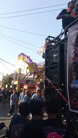 A street procession is underway, indicated by a crowd of people walking along a paved road lined with buildings. A large, elaborate float, resembling a mythical creature with white, yellow, and purple decorations, is the central focus. It is adorned with what appears to be shimmering strands and possibly lights. To the right, a large speaker system is set up, mounted on a sturdy structure, likely part of the procession's sound amplification. The sky is clear and blue, suggesting daytime. Some individuals are looking towards the float, while others are engaged in conversation or walking. The overall atmosphere suggests a cultural event or celebration.