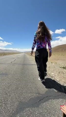 The video shows a person walking away from the camera along a long, straight asphalt road under a bright blue sky with scattered white clouds. The setting appears to be a desolate, arid landscape with low, rocky hills and distant mountains, consistent with Mainpu township, China.

The person is wearing a long-sleeved, colorful top with a blue, purple, and pink tie-dye pattern, loose-fitting black pants, and white sneakers. Their long, dark hair flows down their back. Long shadows are cast on the road, indicating it is daytime and sunny.

As the person walks, a black SUV drives past them in the opposite direction on the left side of the road at the 0:02 mark. Further down the road, another vehicle is visible in the distance. To the right of the road, power lines and utility poles can be seen extending into the landscape. The road has faint white and yellow markings. The overall scene suggests a remote, open highway journey.
