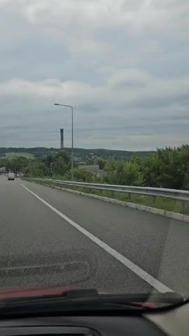 The video shows a view from inside a moving vehicle, looking out onto a road. The road appears to be a highway or a major road with multiple lanes and a guardrail on the right side. The vehicle is traveling on the right side of the road. The landscape outside is rural, with green hills, trees, and some buildings in the distance. There is a tall, smokestack-like structure visible in the far distance, which appears to be part of an industrial facility. The sky is overcast with gray clouds. There are a few other vehicles visible on the road ahead, and some smaller buildings and scattered trees along the roadside. The video appears to be captured during daytime.
