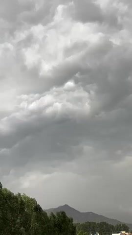The video shows a time-lapse of a stormy sky. Dark, heavy clouds move across the frame, obscuring the sun. In the foreground, the tops of green trees are visible, their branches bending in the wind. In the distance, mountains can be seen beneath the stormy sky. The overall impression is one of an approaching storm.