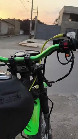 A low-angle shot captures the view from the handlebars of a green and black motorcycle. The handlebars are painted bright green, and the motorcycle's gas tank is matte black. Wires and cables are visibly connected to the handlebars. A hand reaches out to twist the throttle. The engine revs. In the background, a street scene unfolds. A dark-colored car passes behind the motorcycle, followed by a silver car. Buildings are visible on either side of the street, silhouetted against a sunset sky.  A utility pole stands near a metal gate. The overall atmosphere is urban.