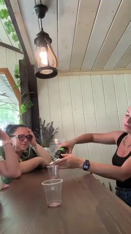 The image captures three women gathered around a dark wooden table in what appears to be a casual indoor or semi-outdoor setting, possibly in Rassvet, Russia. The background features light-colored wooden paneling, a rustic hanging lantern with an exposed bulb, and decorative artificial ivy draped along a beam.

In the foreground, a woman on the right, wearing a black tank top and a smartwatch on her left wrist, is actively pouring liquid from a green bottle into small, clear plastic cups. Her arm displays visible tattoos.

Two other women are seated at the table, facing the camera. The woman in the center wears a white top, a white bandana, and sunglasses, resting her chin on her hand with a slightly pouting expression. To her left, another woman wears a brown hat and sunglasses, also resting her chin on her hand with a similar pose. Further back on the left, a third woman with glasses and dark hair, wearing a green top, is visible, resting her hand on her head, appearing contemplative or relaxed.

On the table are several empty and partially filled clear plastic cups. A small pot containing dried lavender or similar decorative plants is visible on a shelf in the background. The overall atmosphere suggests a relaxed social gathering or tasting event. Natural light appears to be entering from a window or opening behind the women.