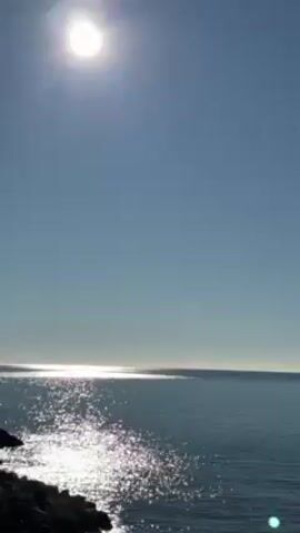The video shows a view of the sea, a rocky coastline, and a town built on a cliffside in Cinque Terre, Italy. The sun shines brightly in a clear blue sky, reflecting on the water's surface. The camera pans up from the sea to reveal a cluster of colorful buildings clinging to the side of a steep cliff, part of the picturesque town. The houses are painted in various pastel shades, and vegetation covers the upper part of the cliff.