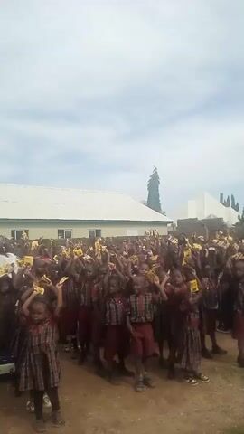 The video captures a large group of young school children standing outdoors, facing forward, and raising small yellow objects above their heads. They are wearing school uniforms consisting of plaid shirts and maroon shorts or skirts. The children appear to be in an open area with a dirt or gravel ground. In the background, there is a building with a white roof and light-colored walls. The sky is overcast. The overall impression is of a school event or activity where the children are participating enthusiastically.