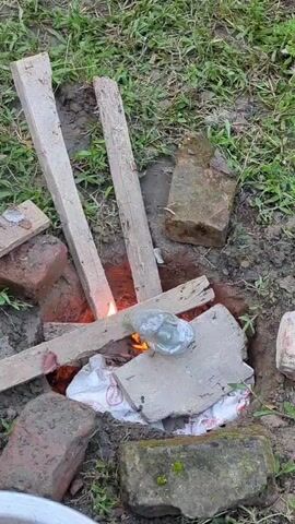 A close-up, high-angle shot shows a small, improvised fire pit dug into the ground. The pit is lined with bricks and contains burning wood, igniting a white plastic bag within. Several pieces of wood are propped up, forming a makeshift support structure over the fire. A silver, flattened object, possibly a metal can or container, is placed on a concrete slab above the flames, appearing to be heated or melted. The surrounding area is covered with dirt and dry grass, with more bricks scattered around the pit.
