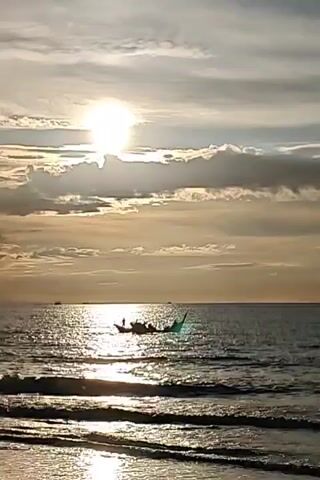 A wide shot of a body of water with a sunset in the background. The sun is a bright orb in the sky, surrounded by clouds. In the middle of the frame, two boats are sailing on the water. The boat on the left is smaller and has one person on it. The boat on the right is larger and appears to have several people on it. The waves are gently rolling towards the shore.
