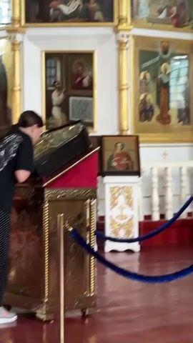 A woman with long brown hair tied in a ponytail, wearing a black t-shirt with silver wings on the back and a long black and white polka dot skirt, stands in an Orthodox church. She is kissing an ornate gold reliquary adorned with red velvet and gold trim, which is placed on a pedestal. The reliquary is open, and the woman is leaning over it. Behind her, a large crucifix hangs on the wall, and several religious icons are displayed. The floor of the church is a deep red. A blue rope barrier separates the viewer from the area around the reliquary. The woman briefly looks up and then returns to kissing the reliquary. She then moves slightly to her left and appears to make the sign of the cross before turning away and walking out of frame.